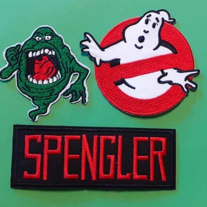 Może przedstawiać: Trzy haftowane łatki z postaciami z filmu Ghostbusters. Pierwsza łatka to zielony duch z czerwonymi ustami. Druga łatka to czerwone koło z przekreśloną przekątną linią i białym duchem w środku. Trzecia łatka jest czarna z napisem "SPENGLER" w kolorze czerwonym.