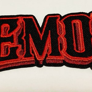 Może przedstawiać: Czerwona naszywka haftowana z napisem "DEMON" w pogrubionym, trójwymiarowym stylu. Litery są obwiedzione czernią i wypełnione czerwienią.