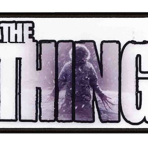 Puede incluir: Un parche blanco con contorno negro que presenta el texto "THE THING" con una foto de una criatura de la película "The Thing" en el fondo.