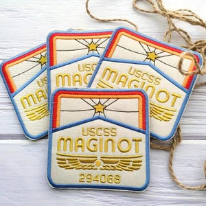 Może przedstawiać: Haftowane naszywki z kremowym tłem, z napisem "USCSS MAGINOT" w kolorze złotym, gwiazdą i wzorem skrzydeł. Naszywki mają niebieską obwódkę i są ozdobione czerwonymi, pomarańczowymi i żółtymi akcentami. Widoczny jest również numer "294068".