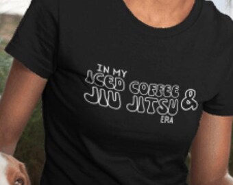 May include: A woman wearing a black t-shirt with the text "In my iced coffee & Jiu Jitsu Era" in white. She is sitting on the ground with a brown and white dog.