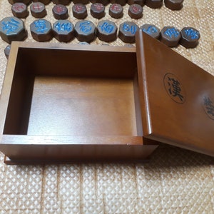 May include: A wooden box with a lid, the lid is open and the box is empty. The box is brown and has Chinese characters on the lid. The box is sitting on a brown surface.