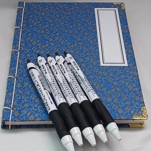 May include: A blue notebook with a gold trim and a white label on the cover. Six black and white pens are lying on top of the notebook.