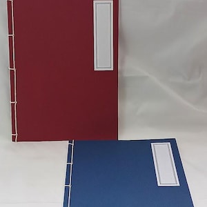 Può includere: Due quaderni vuoti con cuciture bianche lungo il dorso. Il quaderno in alto è rosso con un'etichetta bianca. Il quaderno in basso è blu con un'etichetta bianca.