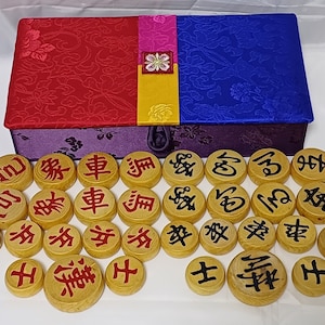 Könnte beinhalten: Ein Satz chinesischer Schachfiguren aus Holz in einer lila, roten, blauen und gelben Schachtel. Die Figuren sind rund und haben chinesische Schriftzeichen in roter und schwarzer Tinte.