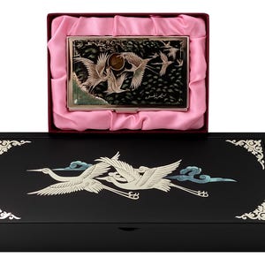 Può includere: Una scatola di legno nera con un coperchio decorativo con un motivo di gru bianco e blu. All'interno, un astuccio argentato con un motivo di gru poggia su raso rosa. L'astuccio è aperto.