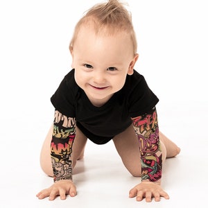 Puede incluir: Un bebé con un body negro con mangas de estampado gráfico de colores está gateando sobre una superficie blanca. El bebé tiene un peinado puntiagudo y está sonriendo.