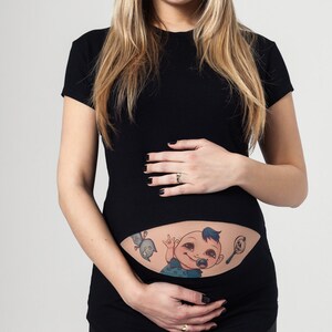K&ouml;nnte beinhalten: Schwarzes Umstandsshirt mit einem Cartoon-Baby-Tattoo-Design. Das Tattoo zeigt ein Baby mit Schnuller, Rassel und Vogel. Die Frau l&auml;chelt und h&auml;lt ihren Bauch.