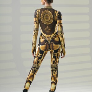 Pu&ograve; includere: Una donna indossa una tuta a stampa oro e nera con un design geometrico. La tuta ha maniche lunghe e un collo alto. La donna indossa stivali neri con plateau.