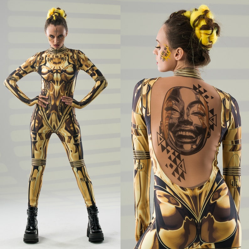 May include: Two women wearing gold and black patterned bodysuits. One woman has a face painted with gold and black geometric shapes. The other woman has a tattoo of a smiling face on her back.