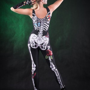 May include: A woman wearing a black skeleton costume with butterflies and roses. The costume is a one-piece jumpsuit with a skeleton design on the front and back. The woman is wearing a flower crown and has a sugar skull makeup design.