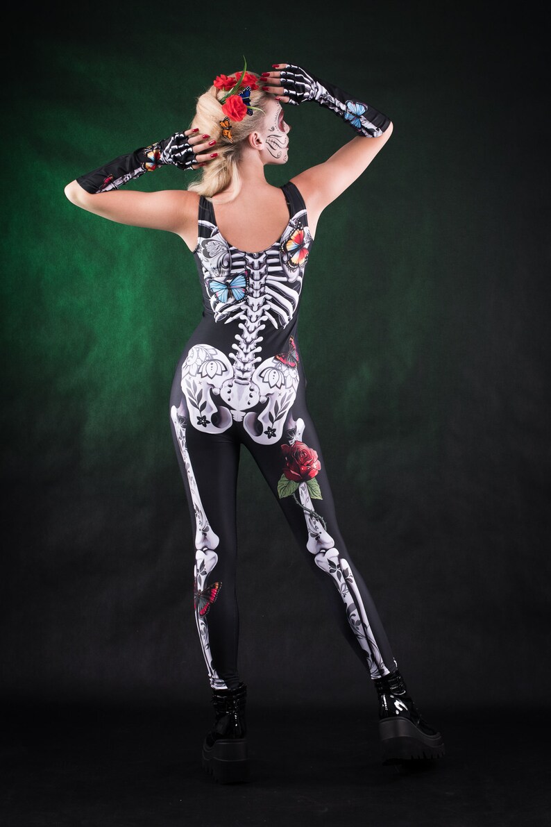 May include: A woman wearing a black skeleton costume with butterflies and roses. The costume is a one-piece jumpsuit with a skeleton design on the front and back. The woman is wearing a flower crown and has a sugar skull makeup design.