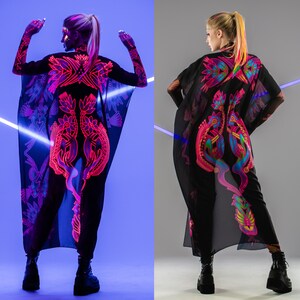 Pode incluir: Duas mulheres vestindo t&uacute;nicas pretas transparentes com padr&otilde;es florais coloridos em neon. As t&uacute;nicas s&atilde;o longas e fluidas, e as mulheres usam cal&ccedil;as pretas e botas.