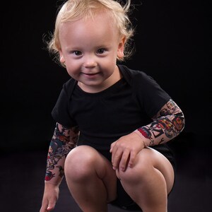 Puede incluir: Un ni&ntilde;o peque&ntilde;o con cabello rubio est&aacute; en cuclillas, usando una camisa negra con mangas de tatuaje coloridas. El ni&ntilde;o sonr&iacute;e y mira a la c&aacute;mara.
