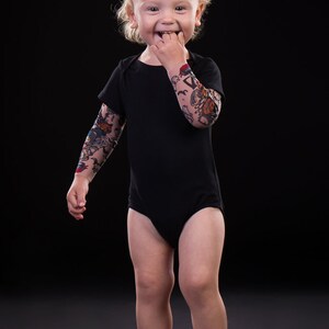 Puede incluir: Un ni&ntilde;o peque&ntilde;o con un body negro y un dise&ntilde;o de tatuaje de manga colorido en su brazo derecho. El ni&ntilde;o sonr&iacute;e y tiene el pelo rubio.