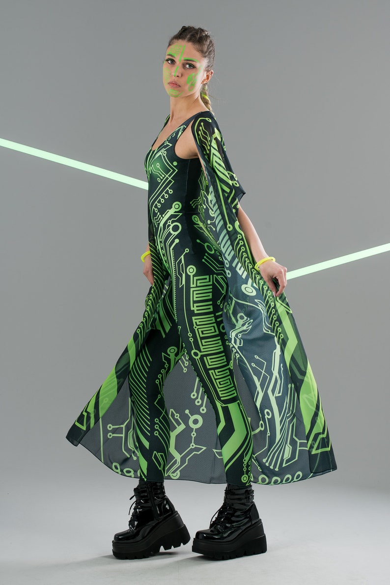 May include: A woman wearing a black and green jumpsuit with a circuit board pattern. She is wearing black platform boots and has green makeup on her face.