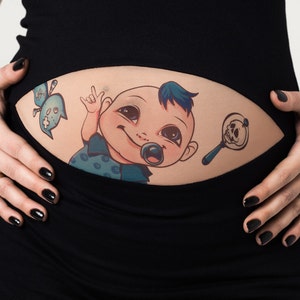 K&ouml;nnte beinhalten: Ein schwarzes Umstandsoberteil mit einem Baby-Motiv. Die Grafik zeigt ein Cartoon-Baby mit Schnuller, blauem Irokesenschnitt und Totenkopf-Rassel. Eine blaue geisterhafte Figur ist ebenfalls enthalten.
