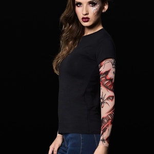 May include: A woman wearing a black t-shirt and blue jeans with a spider web and spider tattoo sleeve on her right arm. She has a spider web design painted on her face.