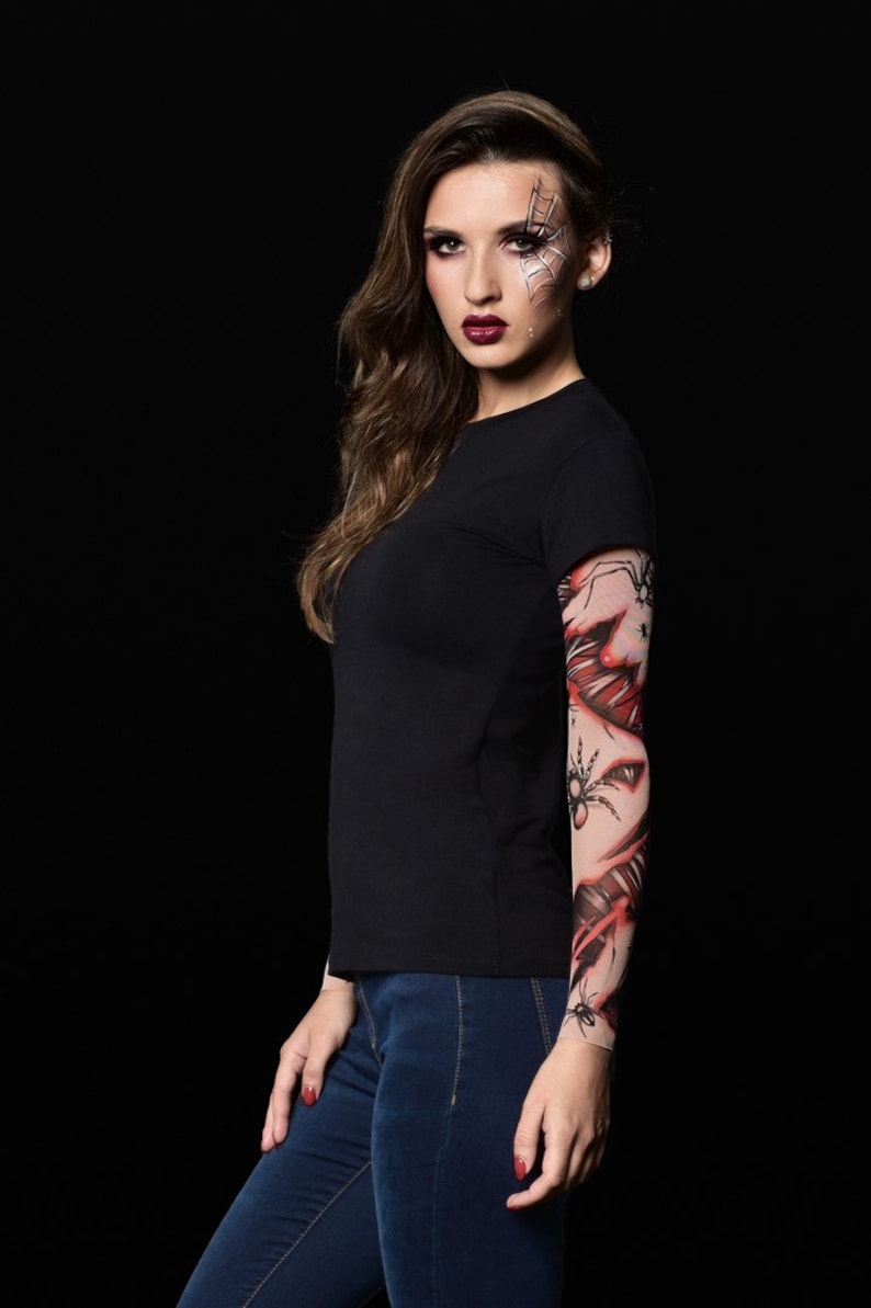 May include: A woman wearing a black t-shirt and blue jeans with a spider web and spider tattoo sleeve on her right arm. She has a spider web design painted on her face.