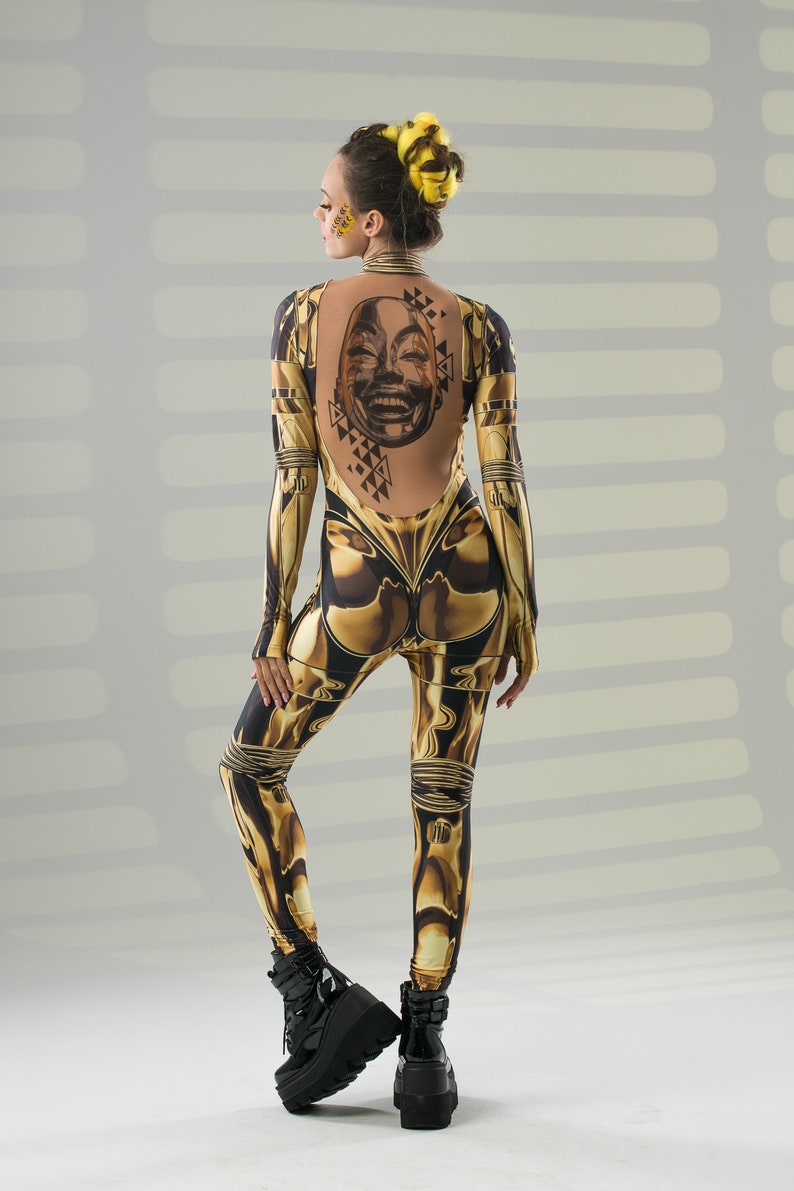 May include: A woman wearing a black and gold jumpsuit with a graphic print of a face on the back. She is wearing black platform boots.