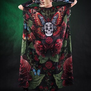 May include: A black sheer maxi dress with a skull and butterfly print. The dress features red roses, green leaves, and a skull with a butterfly on its head. The dress is worn by a person with blonde hair and a flower crown.