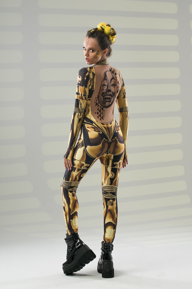May include: A woman wearing a gold and black patterned jumpsuit with a large graphic print on the back. She has yellow hair and is wearing black platform boots.