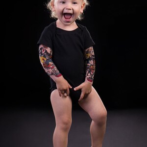 Puede incluir: Un ni&ntilde;o peque&ntilde;o con un body negro y mangas de tatuaje de colores sonr&iacute;e y mira a la c&aacute;mara.