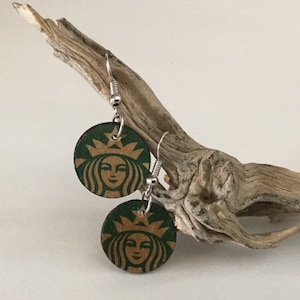 May include: A pair of green and gold earrings with the Starbucks logo. The earrings are hanging from a piece of driftwood.