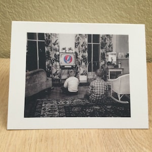 May include: Black and white photograph of two children sitting on a patterned rug, watching a television. The TV displays a colorful Steal Your Face logo. The room has floral curtains and a vintage aesthetic.