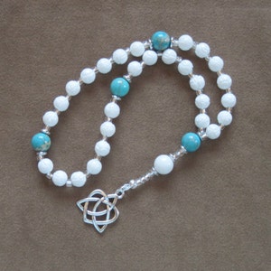 Puede incluir: Mala de oración con cuentas blancas y turquesa con un dije de nudo celta plateado.