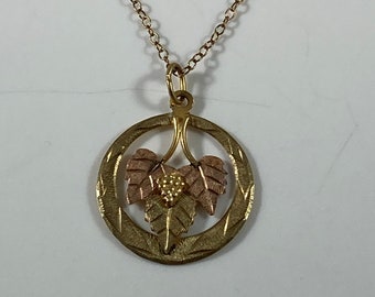 May include: Gold-tone necklace with a circular pendant featuring a grape and leaf design. The pendant has a textured gold outer ring with a cluster of grapes and leaves in shades of gold and rose gold. The chain is delicate.