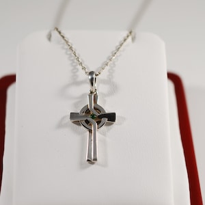 May include: A silver-toned Celtic cross pendant necklace. The cross features a central green gemstone and a detailed design. The necklace is displayed on a white surface with a red border, showcasing the jewelry's craftsmanship.