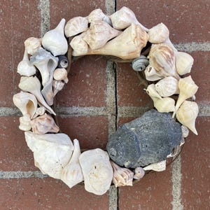 May include: A circular wreath composed of various seashells in shades of white, beige, and gray. The wreath is arranged on a brown base and includes a large, dark gray rock. The background is a red brick wall.
