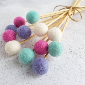 May include: A bouquet of felt balls on wooden skewers, with colours including pink, white, light blue, and purple. The balls are tied together with twine.