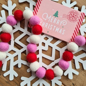 May include: A garland of pink, white and red felt balls on a white snowflake background. The garland is in the shape of a heart. A pink card with the text "Merry Christmas" is in the background.