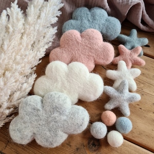 May include: A set of felt cloud and star decorations in shades of grey, pink, and white. The decorations are arranged on a wooden surface with a dried pampas grass stem in the background.