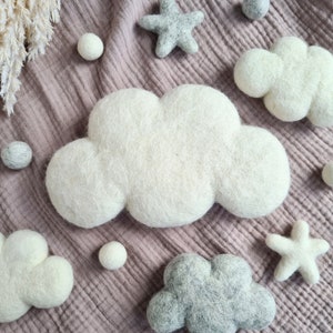 May include: A set of felt cloud and star decorations in white and grey. The clouds are fluffy and the stars have five points. The decorations are arranged on a textured pink fabric background.