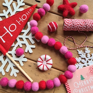 May include: A red felt banner with the text "Ho Ho Ho" is displayed next to a white snowflake. A garland of pink and red felt balls is wrapped around a wooden surface. The garland is decorated with a red felt star and a candy cane.
