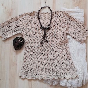 Puede incluir: Un top de ganchillo beige con dobladillo y mangas festoneadas, combinado con un cinturón de cuero marrón y un collar largo marrón con cuentas decorativas. También se incluye una falda de encaje blanco.