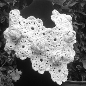 May include: A white crochet shawl featuring a series of interconnected circular floral motifs and three-dimensional rose-shaped embellishments. The garment is constructed with intricate yarn stitching, creating a textured, openwork pattern throughout the piece.