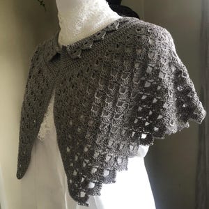 Op de afbeelding: Een grijze gehaakte sjaal met een gegolfde rand. De sjaal is gemaakt met een delicate opengewerkte steek.