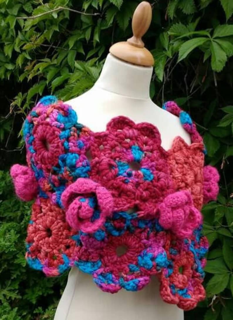 May include: A handmade crochet shawl featuring a vibrant pattern of interconnected floral motifs in shades of bright pink, magenta, and electric blue. The garment is constructed with thick yarn, showcasing textured three-dimensional rose accents.
