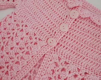 May include: A light pink, handmade crochet cardigan with a scalloped collar and two matching buttons. The openwork design and delicate stitching create a soft, textured appearance. This baby or toddler sweater is a charming addition to any child's wardrobe.