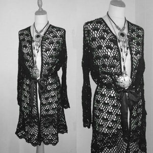 Puede incluir: Un cárdigan de crochet negro con una abertura larga en la parte delantera y un cinturón de lazo. El cárdigan tiene un delicado patrón de encaje y está hecho de una tela ligera y transpirable.