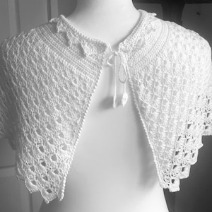 May include: A white, open-front crochet shawl with a delicate, lacy pattern. The shawl features a scalloped edge and a tie closure at the neckline. The intricate design and soft texture create an elegant, vintage-inspired accessory.