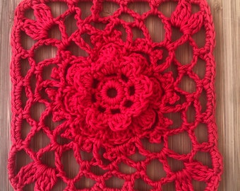 May include: A square, handmade, red crochet doily. The center features a raised floral design, surrounded by an openwork pattern. The edges are finished with a scalloped border. The doily is displayed on a wooden surface.