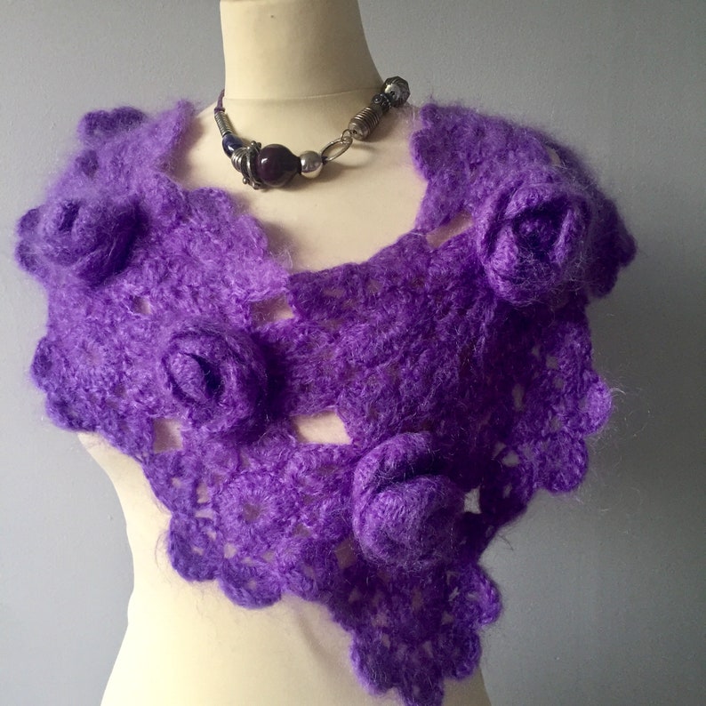 May include: A vibrant purple crochet shawl featuring a delicate openwork pattern and three-dimensional floral embellishments. The shawl is draped over a mannequin and is paired with a beaded necklace featuring dark purple and silver-toned beads.