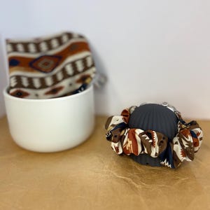 May include: A fabric scrunchie featuring a geometric pattern in shades of brown, cream, navy blue, and orange, wrapped around a dark gray ribbed stand, with a matching fabric piece visible inside a white ceramic bowl in the background.