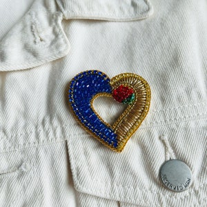May include: A heart-shaped brooch with blue, gold, and red beadwork. The brooch is on a white denim jacket. The heart design features blue beads on one side, gold beads on the other, and a red rose in the center.