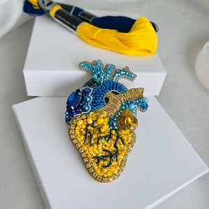 May include: A handcrafted, beaded heart-shaped brooch in shades of blue and yellow, with gold accents. The brooch is embellished with various beads and crystals. A skein of yellow and blue embroidery floss is in the background.
